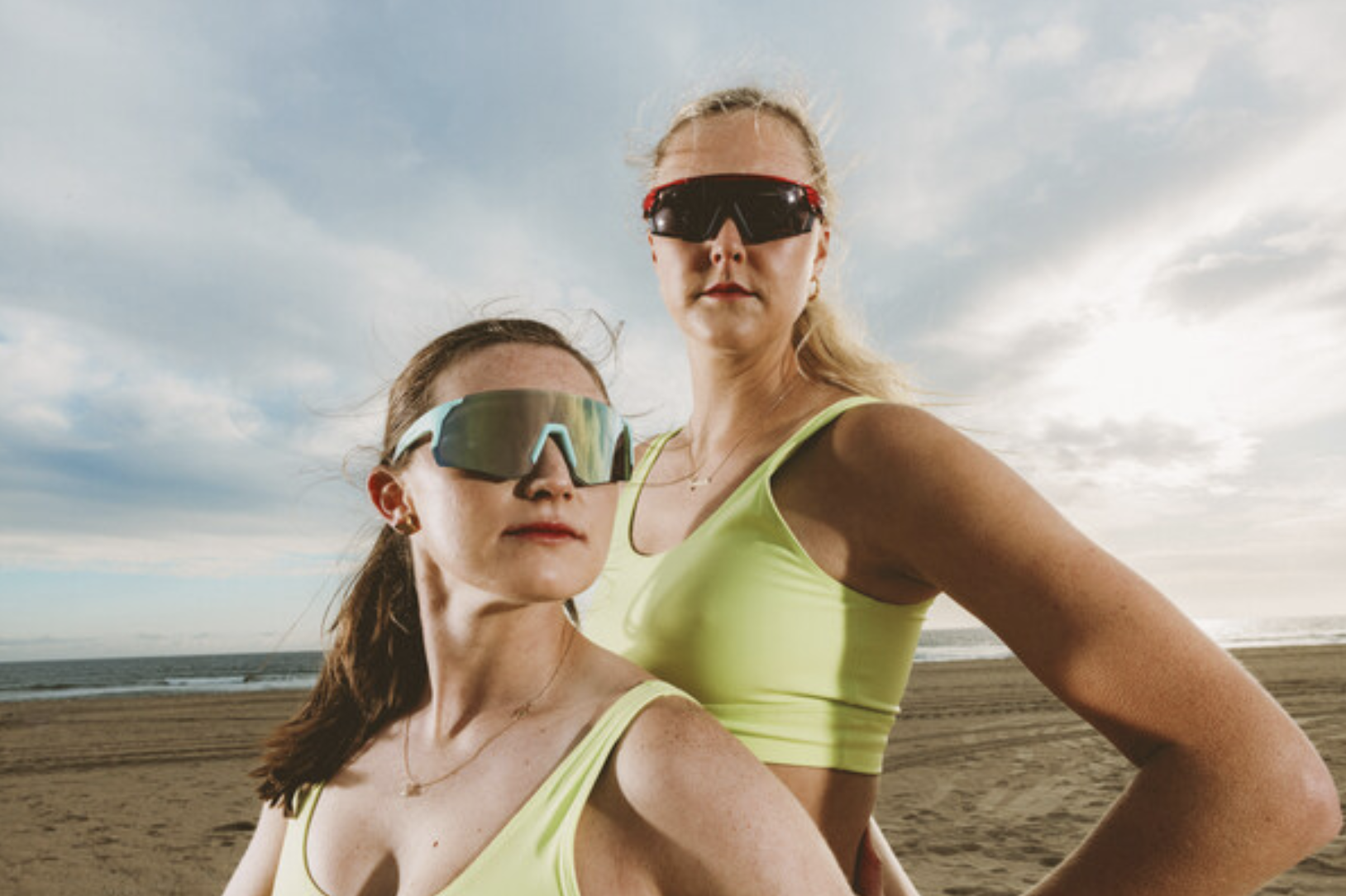 Two women wearing oversized sporty sunglasses and neon sports bras at the beach